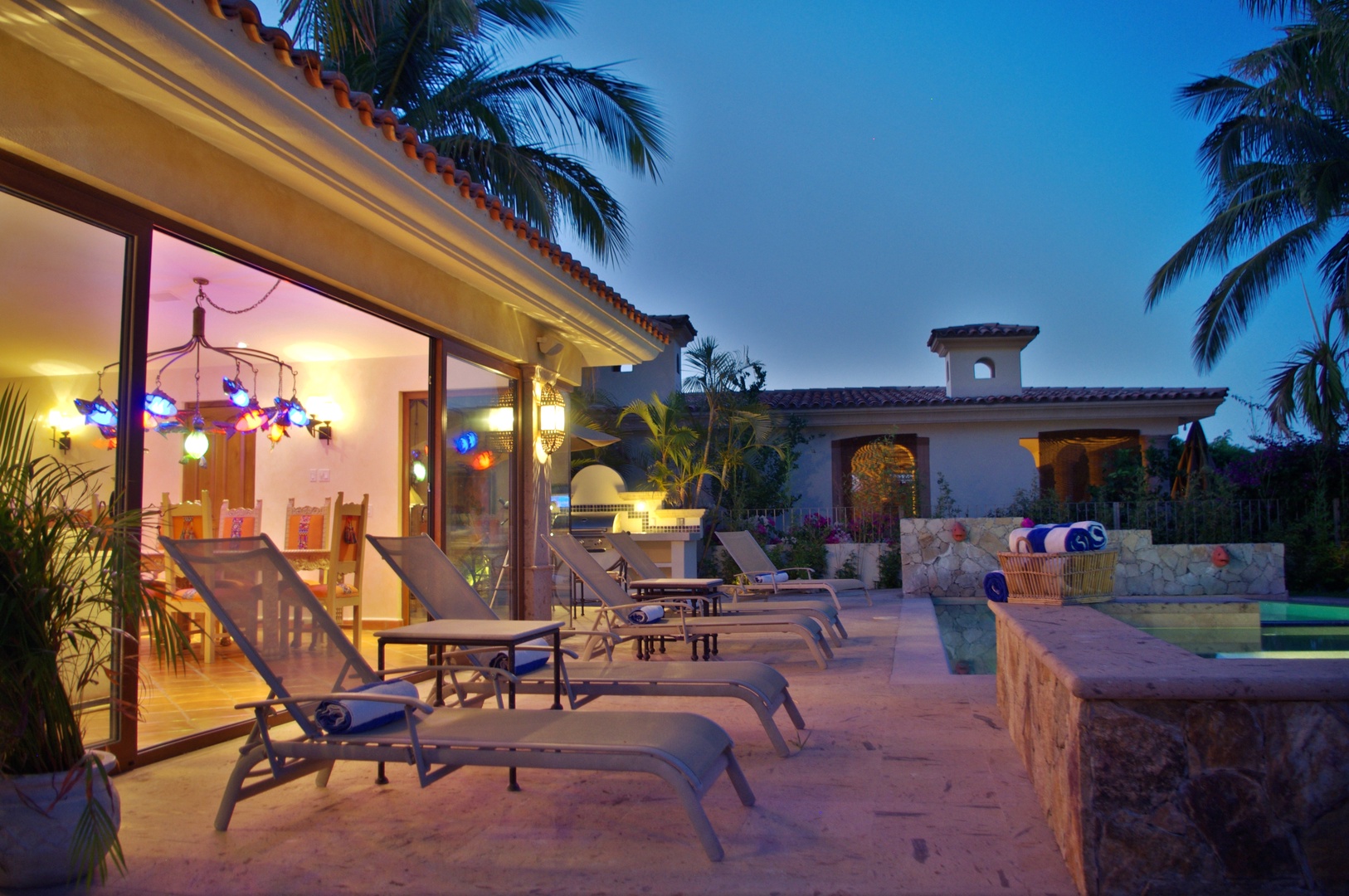 Tropical villa exterior with terrace and pool area at dusk, framed by palm trees and warm ambient lighting throughout the property.
