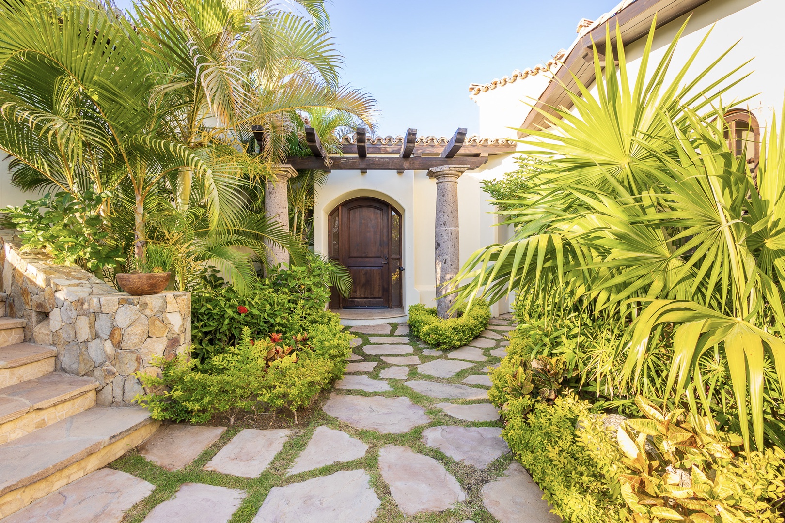 The private entrance features a lush tropical pathway with vibrant palms guiding you beneath a charming pergola to the arched wooden door of this Spanish-style villa.