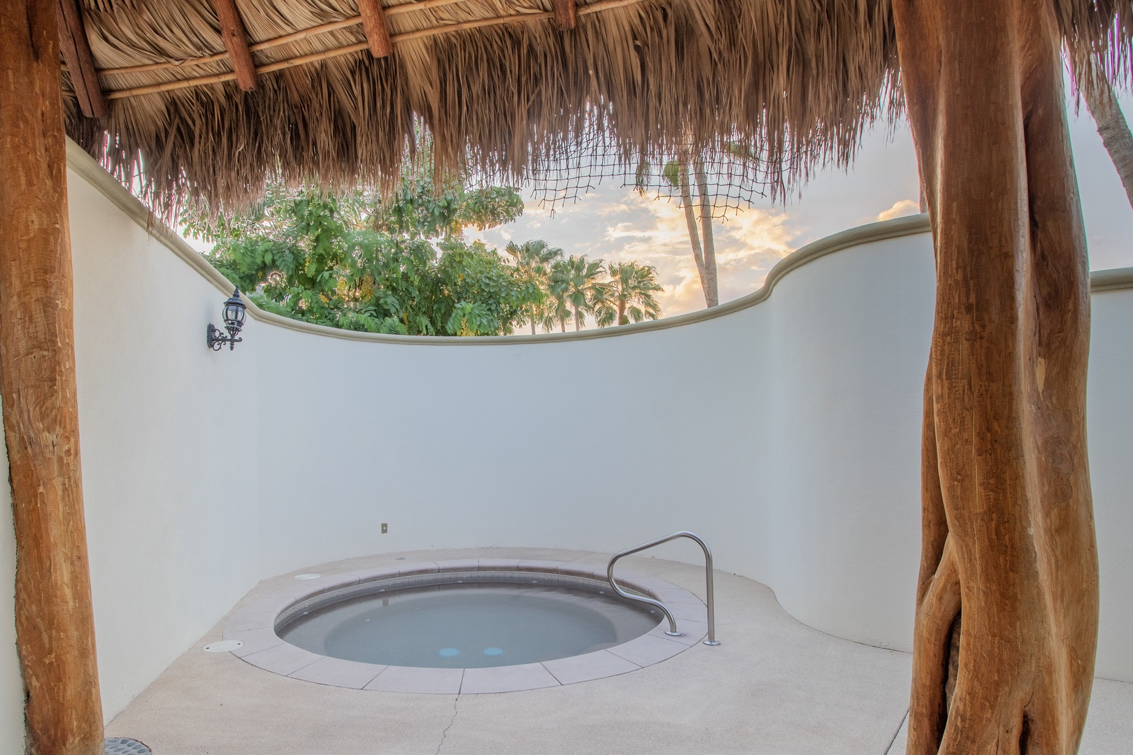 A private hot tub sheltered by a rustic palapa roof, where you can soak under the open sky and glimpse palm trees beyond the curved wall.