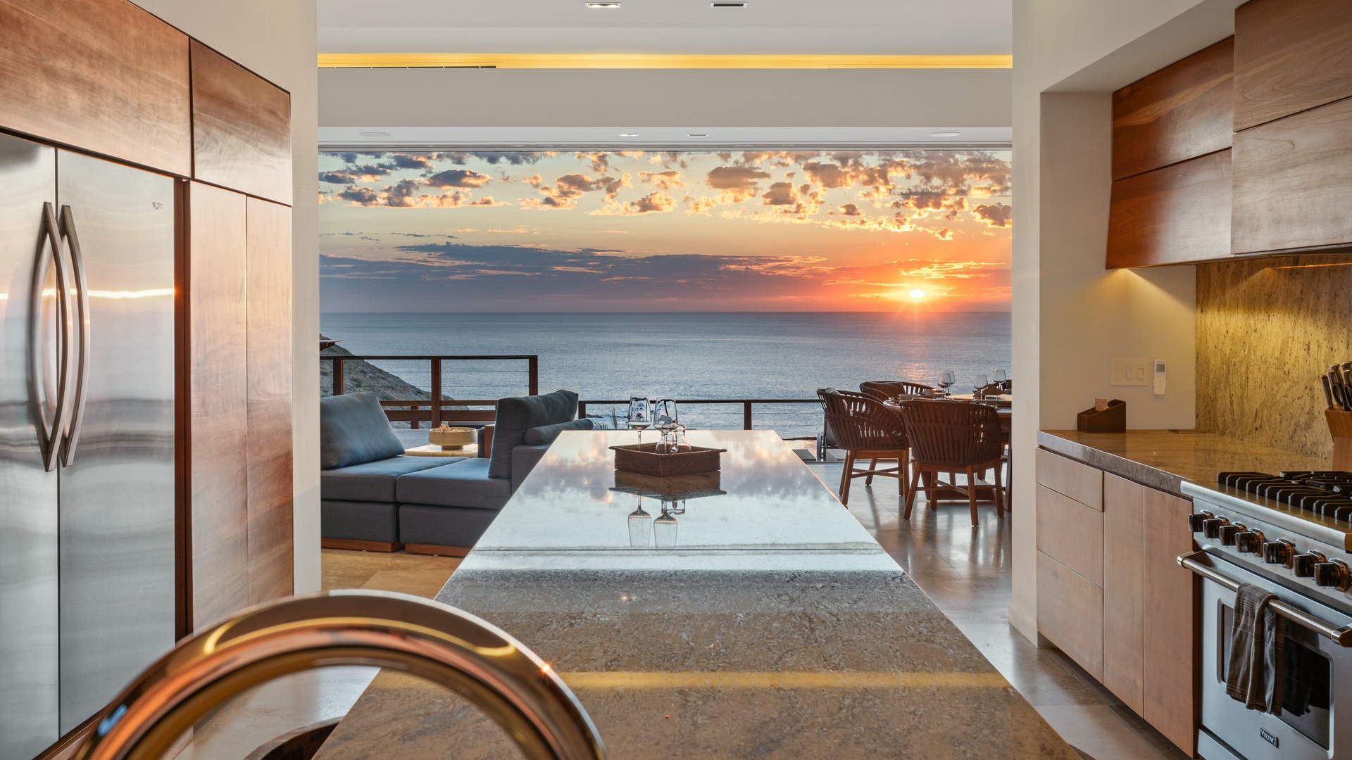 Modern open kitchen seamlessly flows into a living and dining area, framing an unobstructed ocean view. The sunset paints the horizon, visible from the surrounding property.