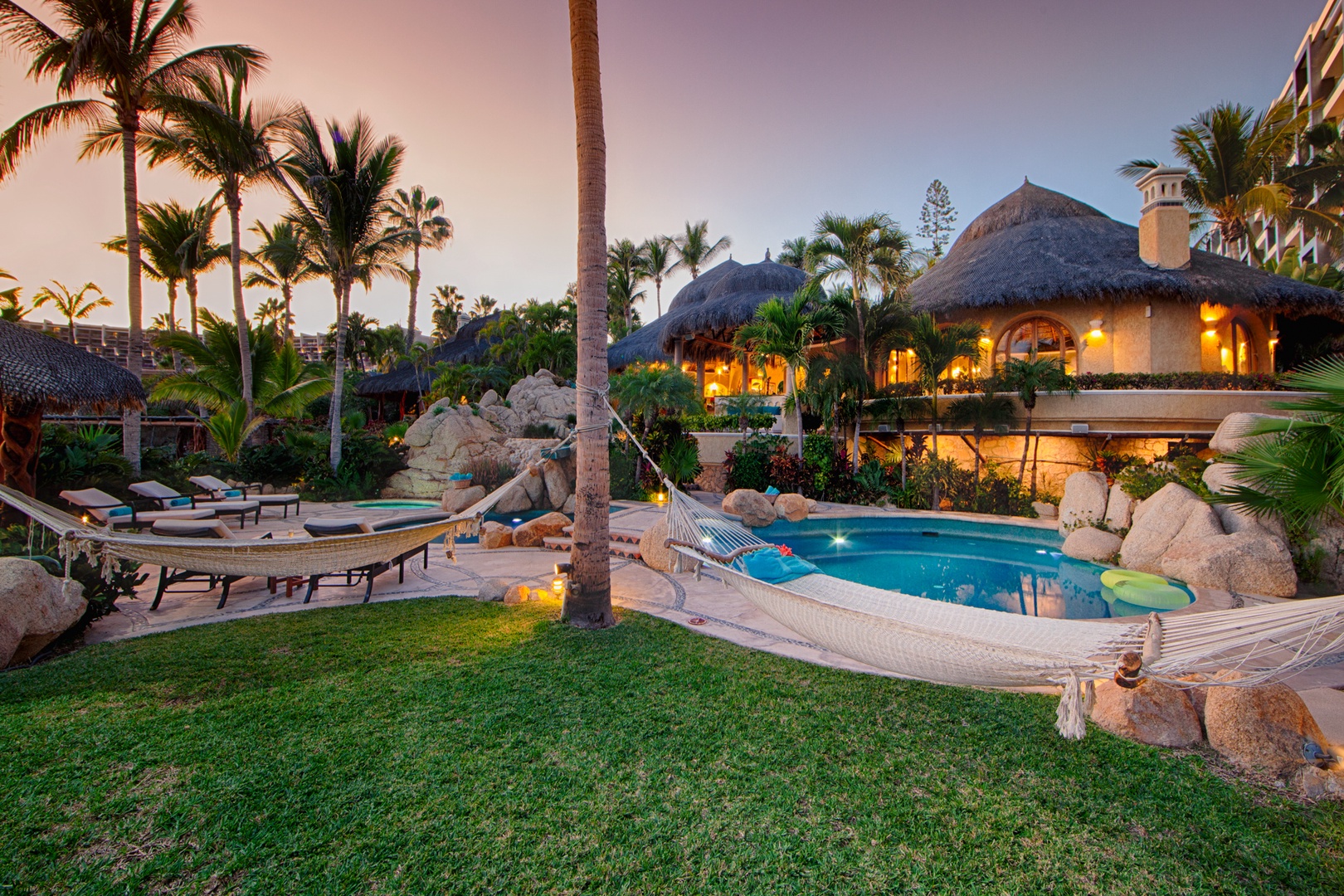 A tropical paradise featuring stunning architecture with thatched roofs, sparkling pool surrounded by palm trees, and magical evening lighting.