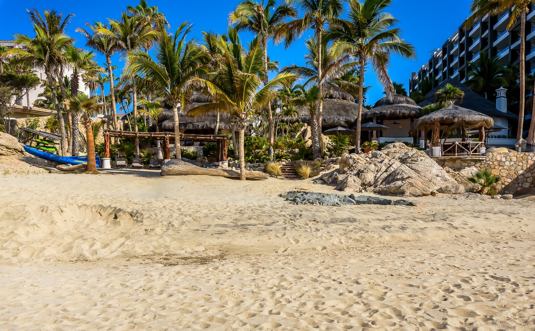 Beachfront property featuring thatched palapas, swaying palms, and soft sandy shores beneath brilliant blue skies—ideal for a sun-soaked coastal escape.