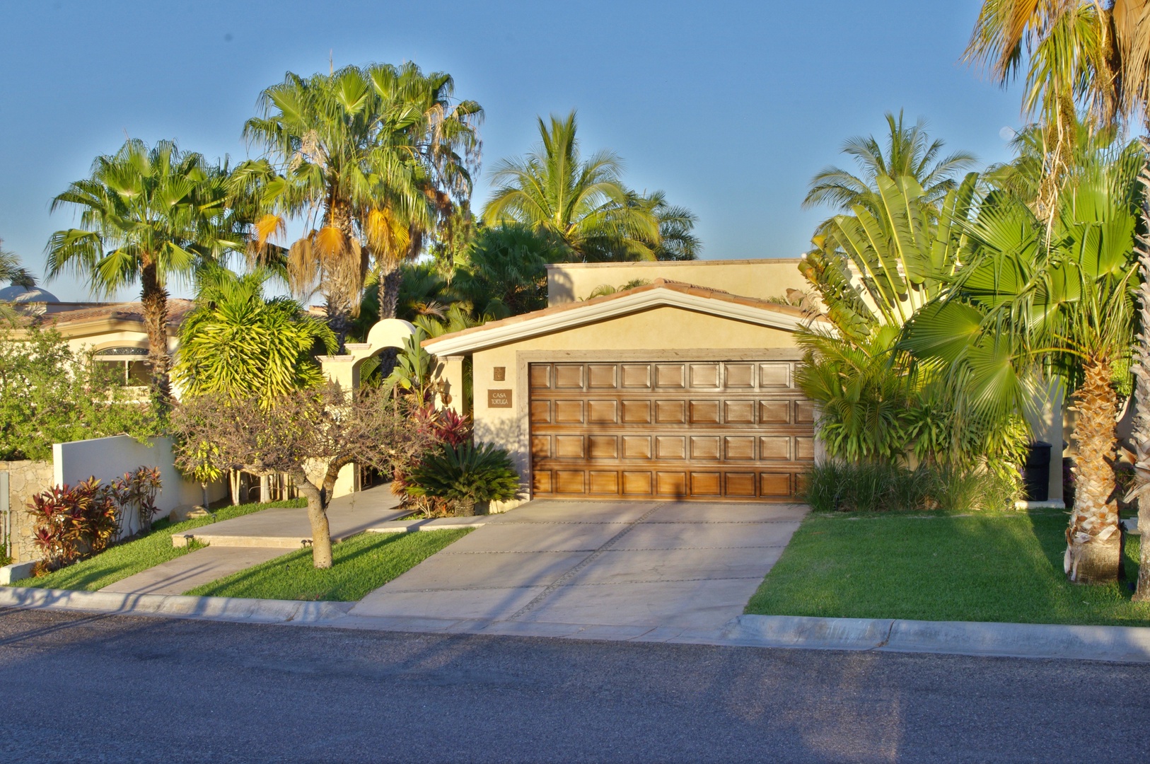 A welcoming single-story retreat surrounded by lush tropical palms and greenery, featuring a two-car garage and private driveway in a tranquil neighborhood setting.