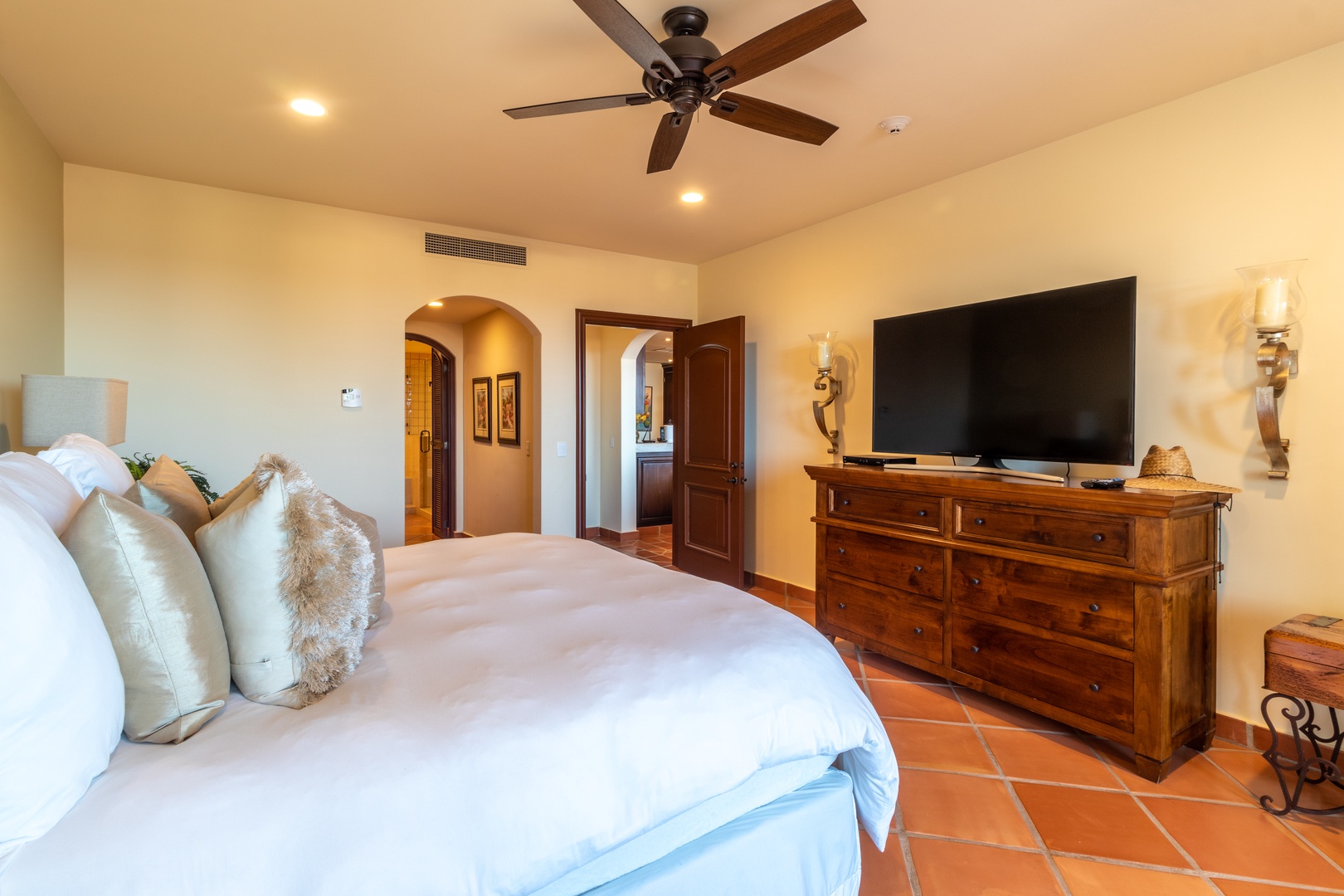Unwind in your serene bedroom featuring a plush bed, rustic wood furnishings, and modern entertainment. The ceiling fan keeps you comfortable while terracotta tiles add authentic charm.