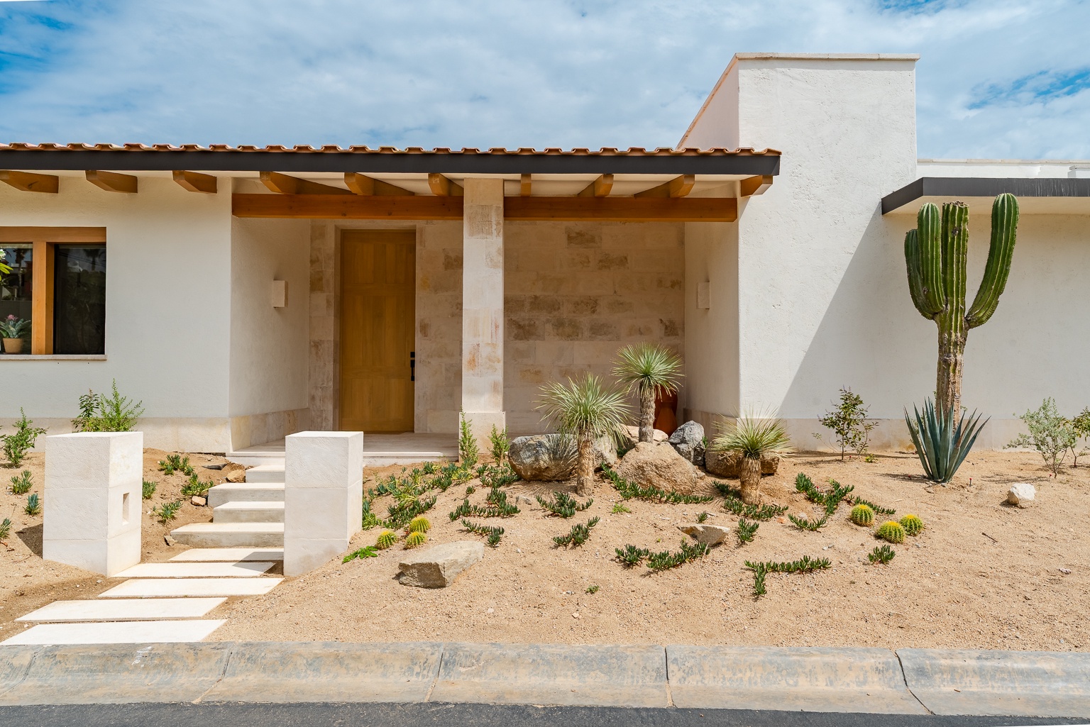 Contemporary desert architecture featuring natural stone accents, exposed wood beams, and native landscaping with mature cacti and agave plants defining the property entrance.