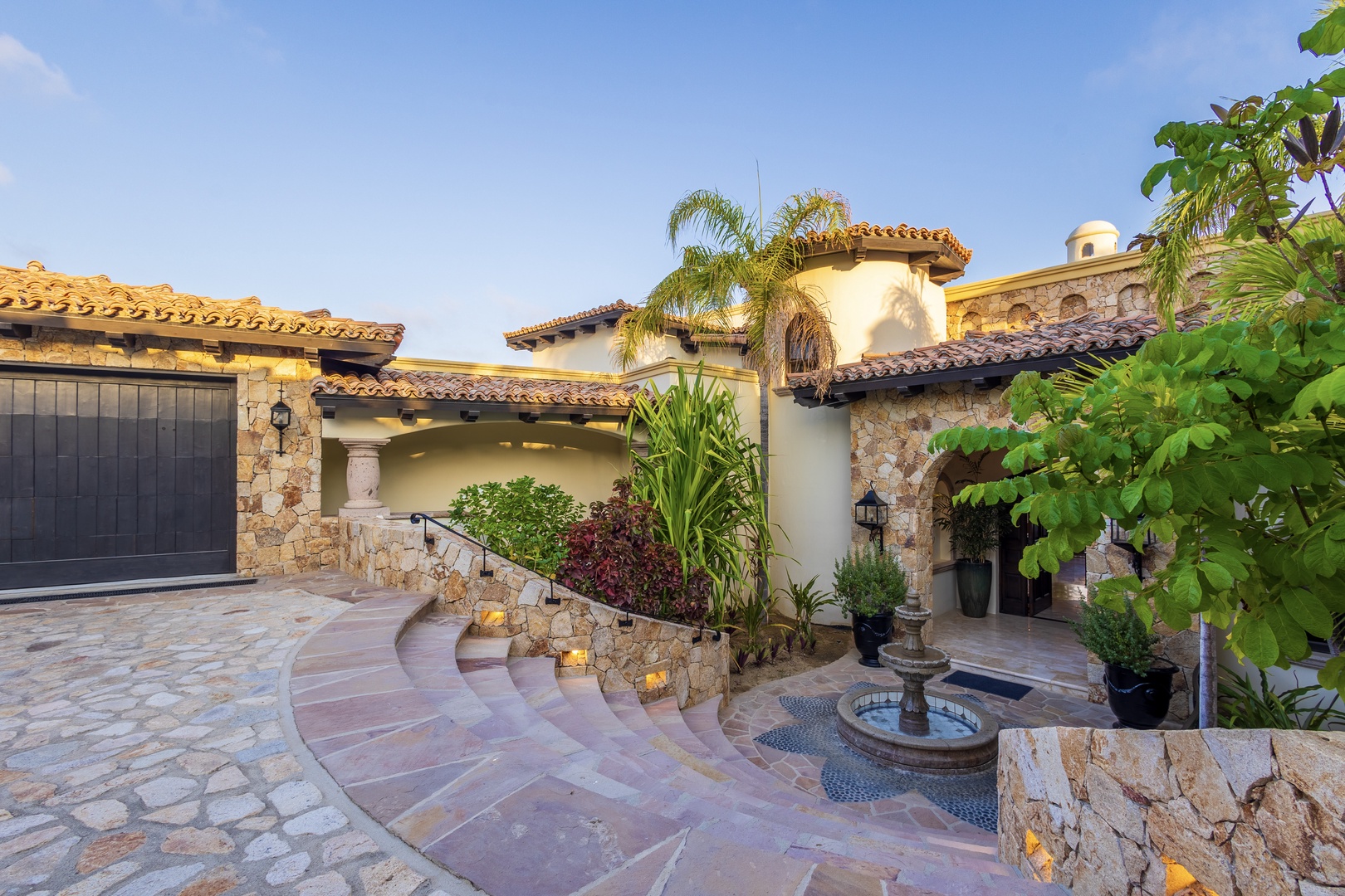 Stunning Mediterranean-style villa exterior featuring stone accents, terracotta roof tiles, lush tropical landscaping, and a decorative courtyard fountain marking the entrance.
