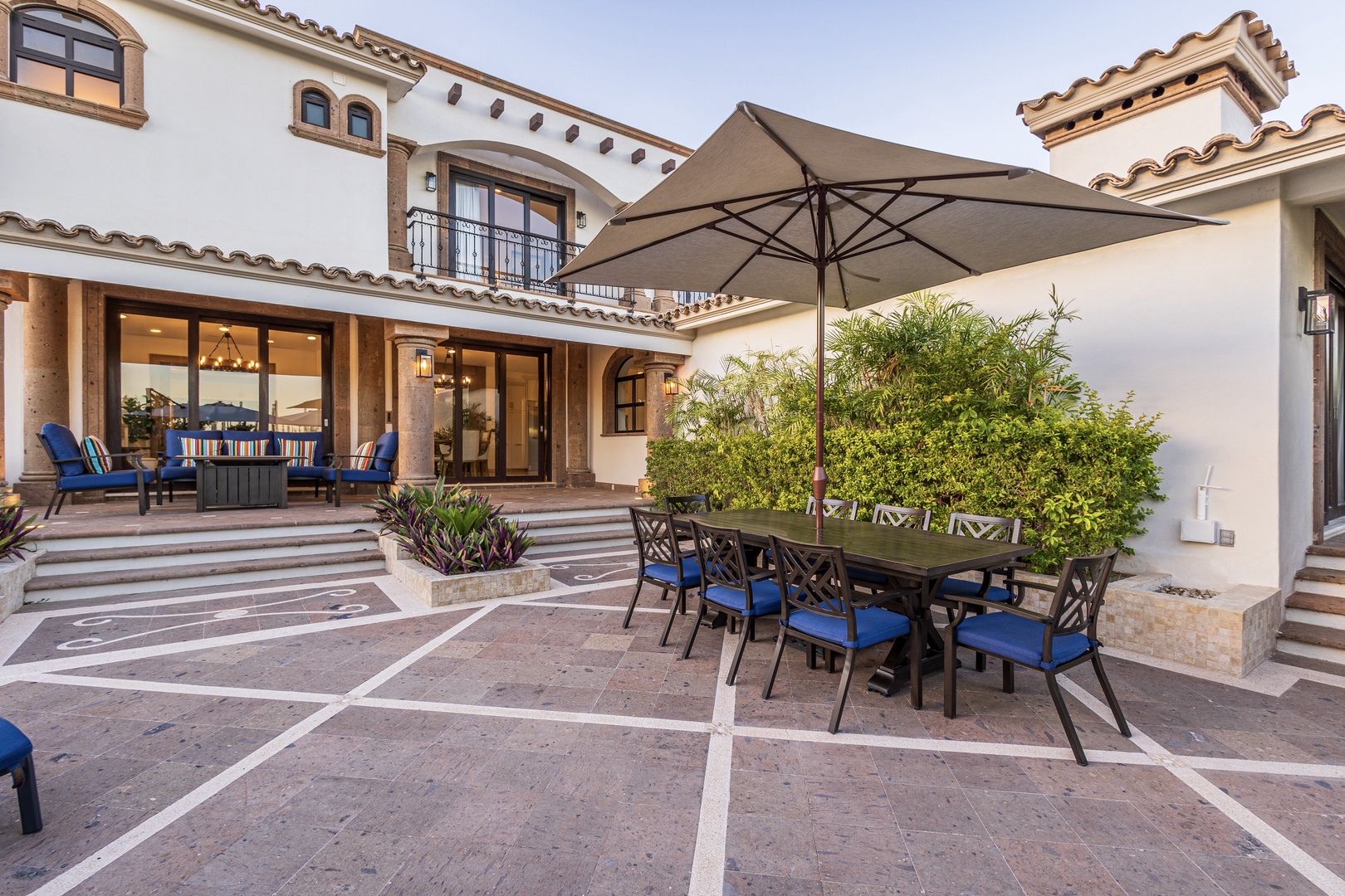A spacious Mediterranean-style villa featuring a private patio with shaded outdoor dining for eight, lush landscaping, and direct access to elegantly lit living spaces.