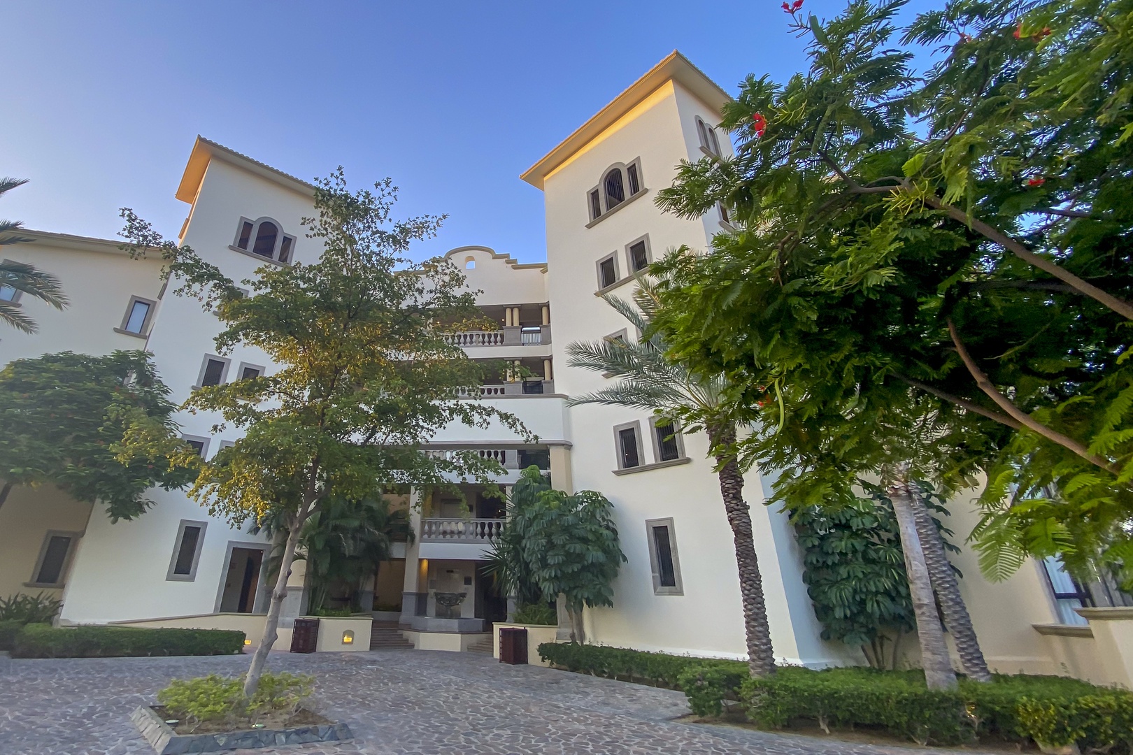 A Mediterranean-style property surrounded by lush palm trees and tropical greenery, featuring elegant white architecture with multiple balconies and a welcoming cobblestone courtyard.