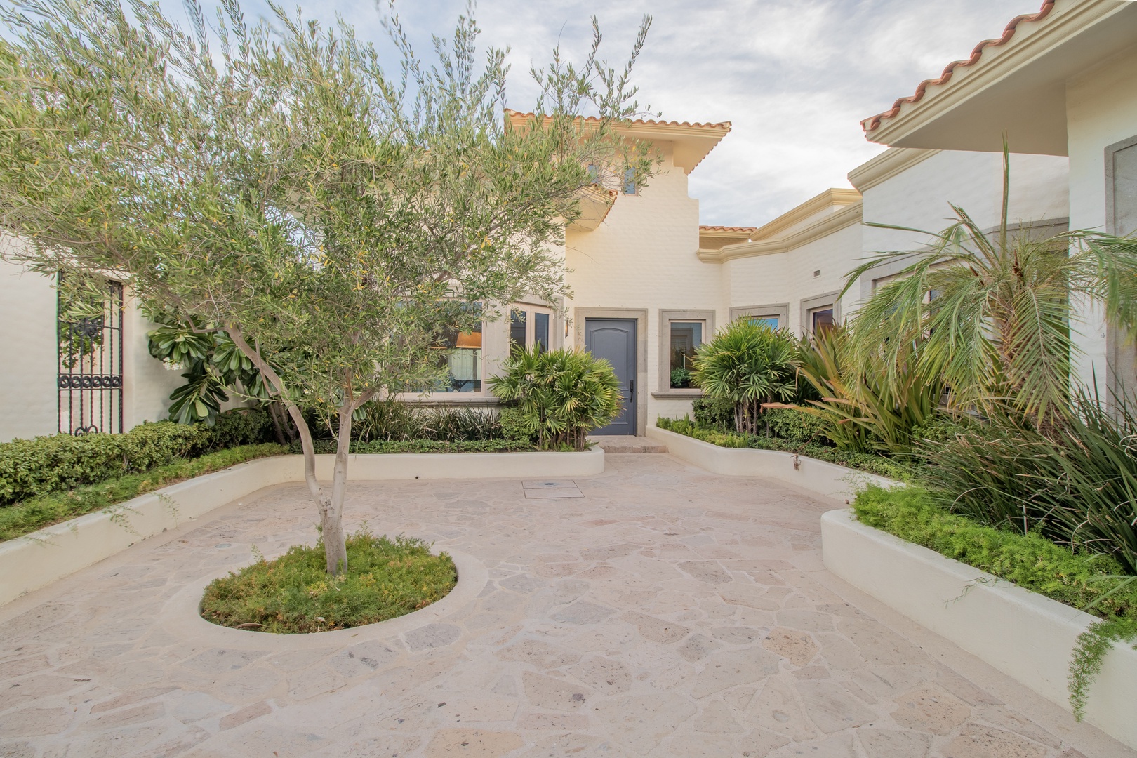 This Mediterranean-style property features a charming courtyard entrance with terra cotta tile roofing, lush tropical landscaping, and distinctive olive trees framing the welcoming facade.