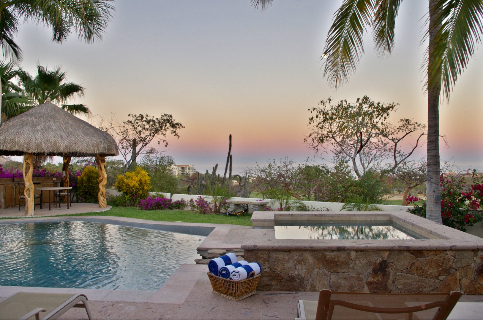 This property offers a serene escape with a swimming pool, hot tub, and tropical gardens set beneath swaying palms and a colorful desert sunset.