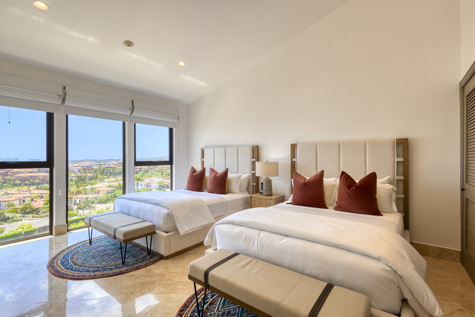Wake up to stunning views from your spacious bedroom with two plush beds dressed in crisp linens and rich rust-toned accents, where floor-to-ceiling windows frame the lush landscape beyond.