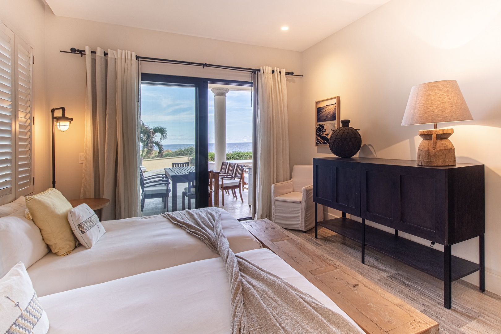 Unwind in your serene coastal retreat featuring twin beds with crisp linens, warm wood floors, and French doors opening to a terrace with stunning ocean views and outdoor dining.