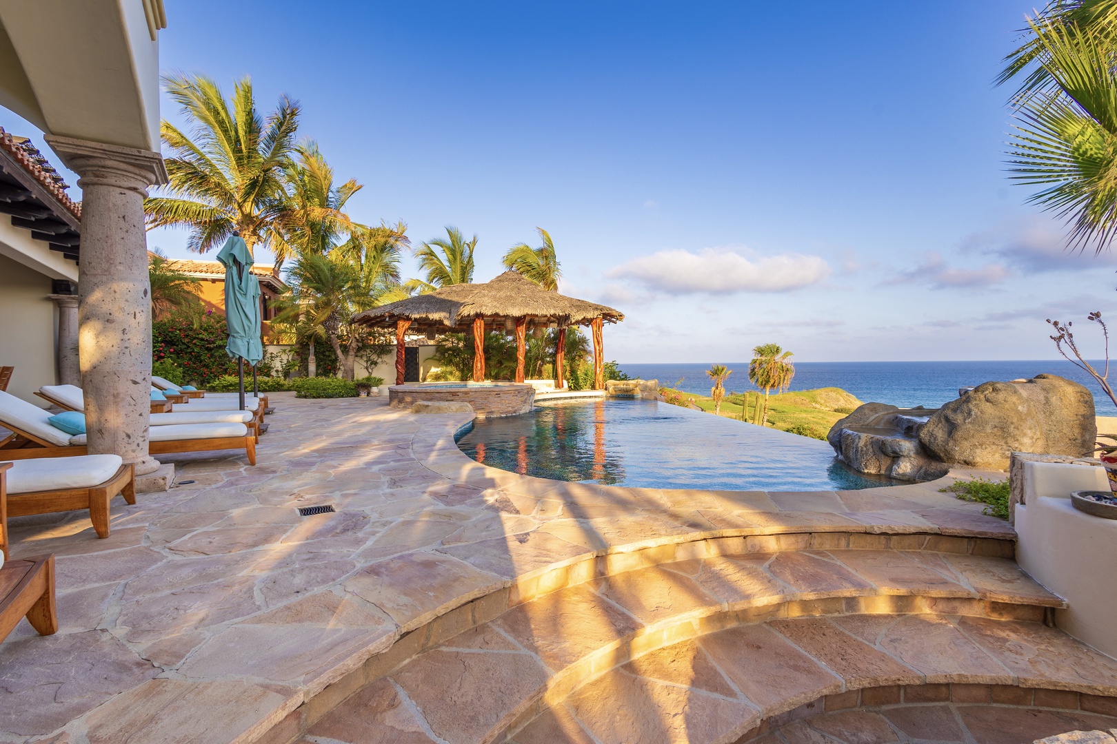 A luxurious infinity pool overlooking the ocean, surrounded by palm trees and a thatched palapa. This coastal oasis features stone decking, lounge chairs, and dramatic natural rock formations at the water's edge.