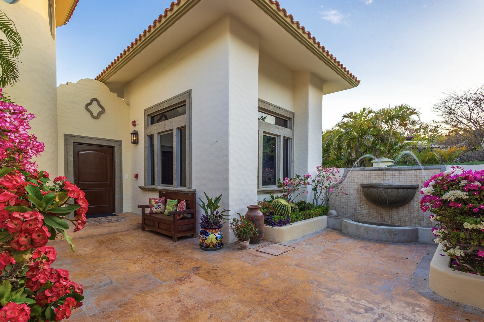Charming villa entrance framed by vibrant bougainvillea blooms and tropical gardens, featuring Spanish tile roof, decorative fountain, and inviting wooden bench by the front door.
