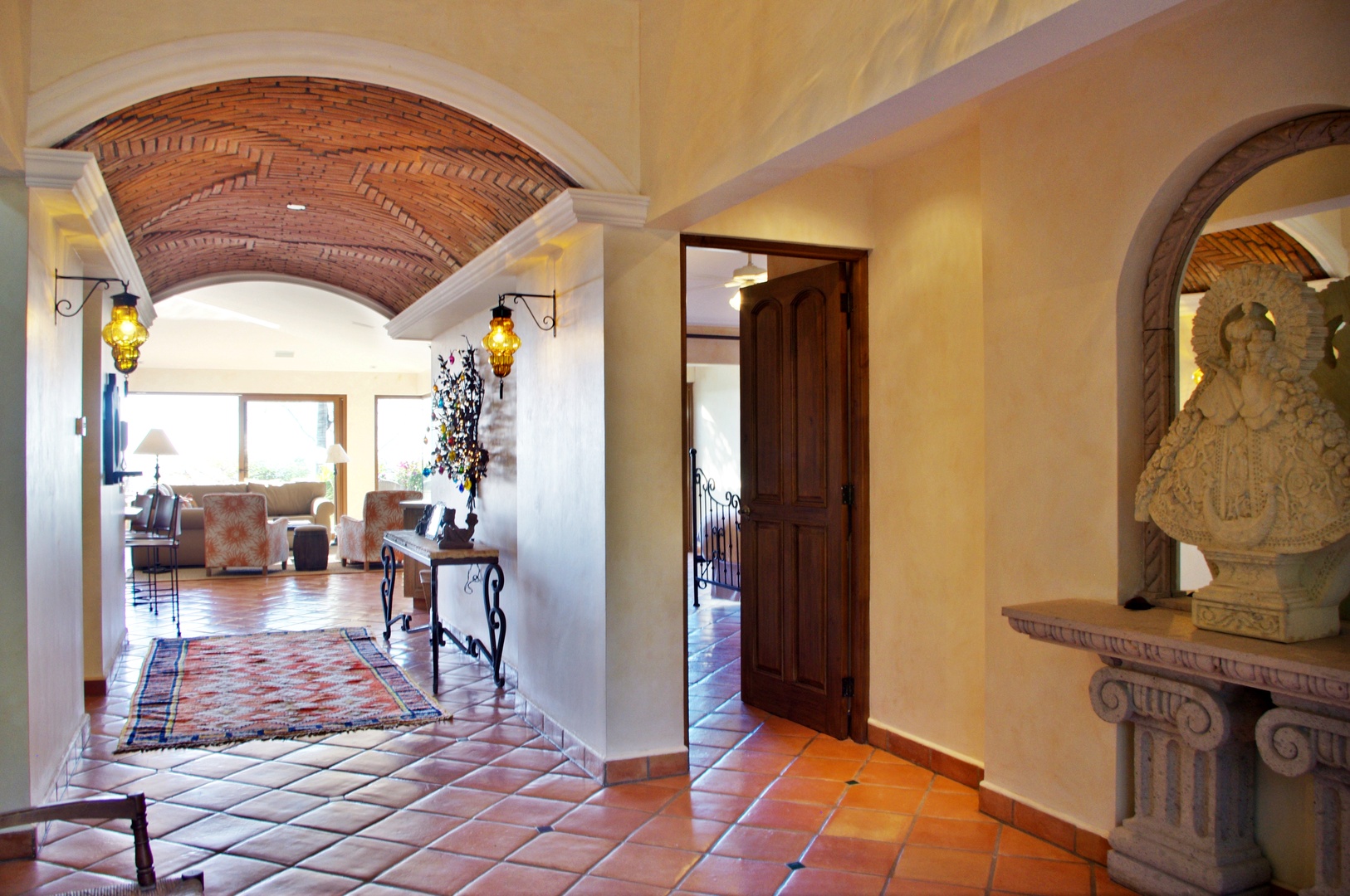 Step through the archway into a sun-warmed hallway where rustic brick ceilings meet terracotta floors, guiding you toward a welcoming lounge bathed in natural light.