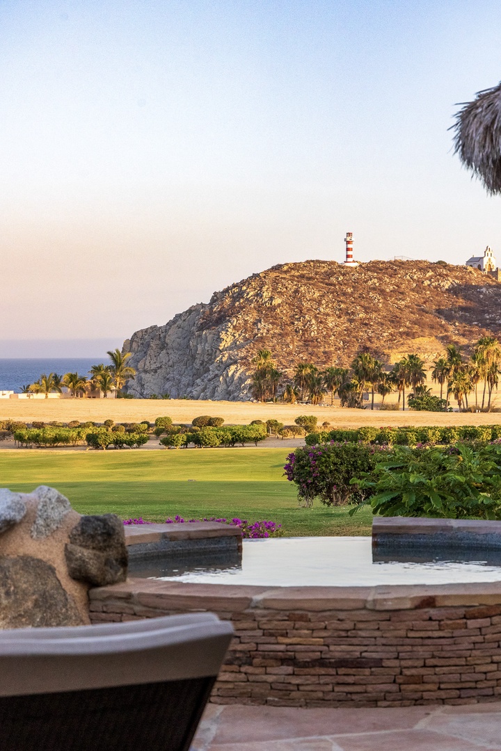 The property grounds overlook manicured lawns, a sandy beach, and a dramatic rocky hillside with a red-and-white striped lighthouse marking the coast.