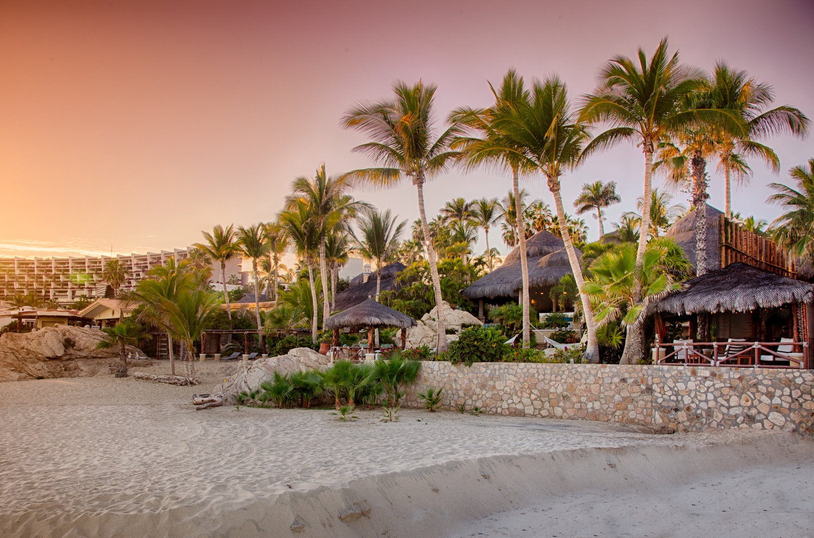 A tropical beachfront resort with thatched-roof palapas and swaying palm trees creates an idyllic sunset scene.
