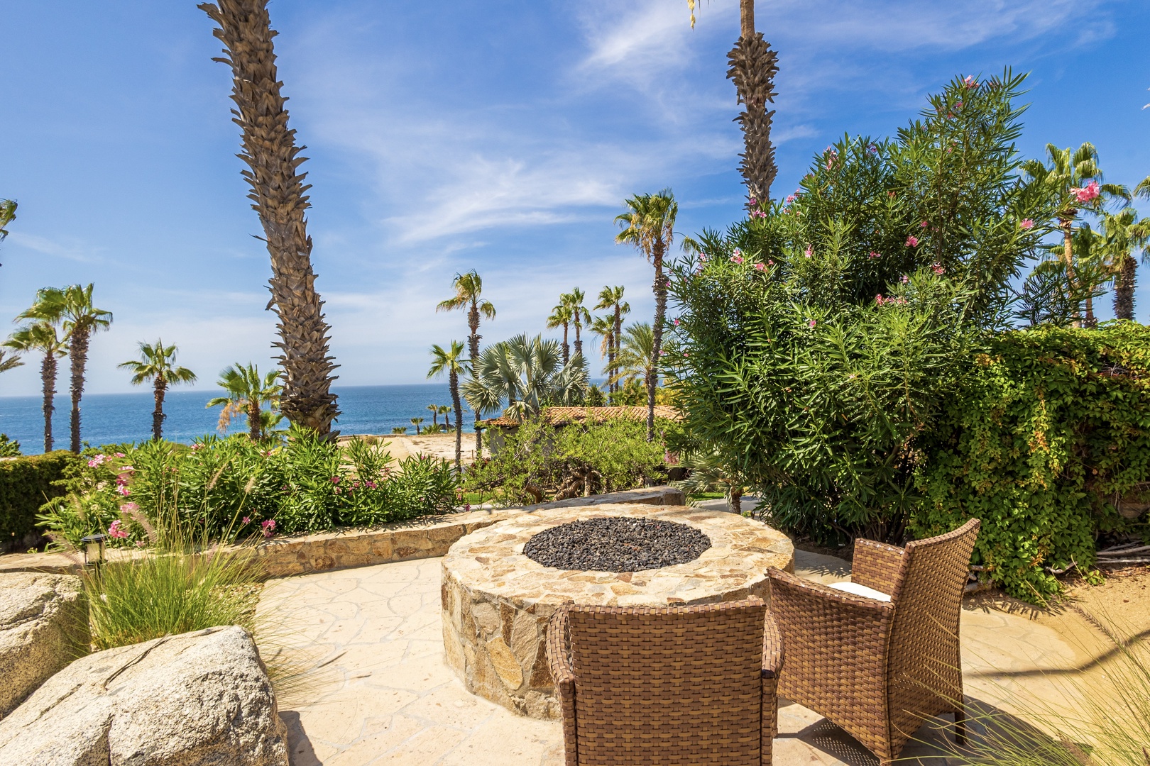 A seaside outdoor patio featuring palm trees, lush tropical landscaping, and a stone fire pit surrounded by woven seating—where ocean breezes and coastal views set the scene for your stay.