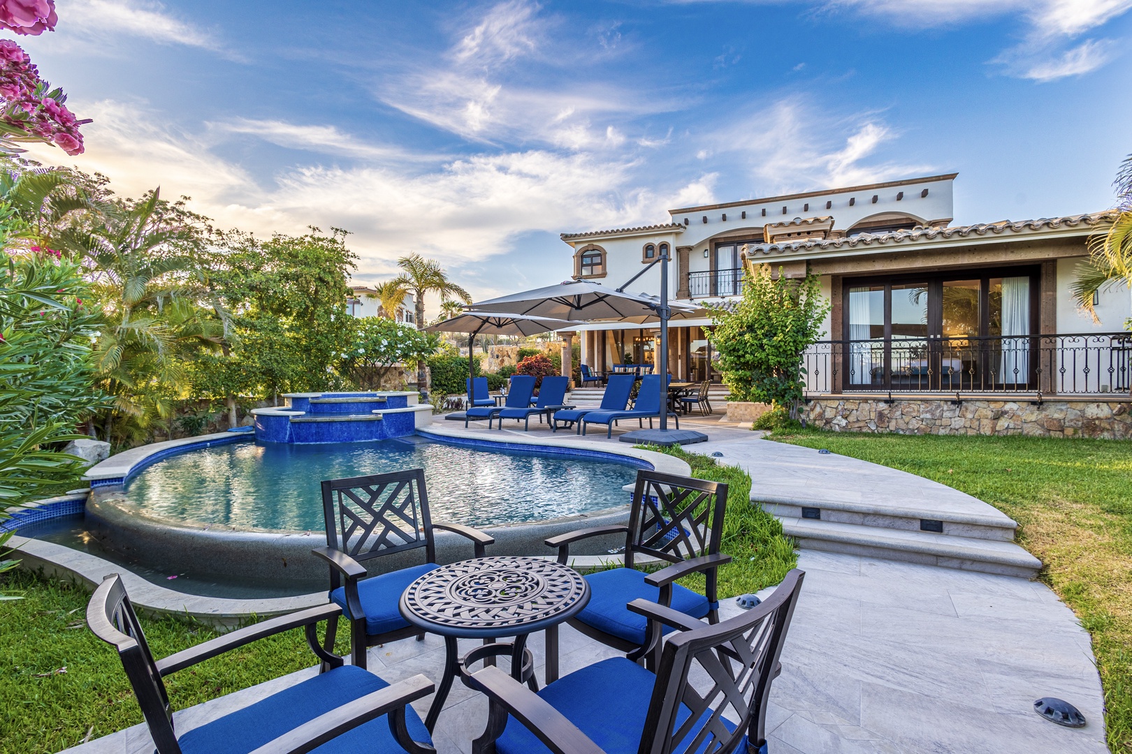 Elegant Spanish-style villa with sparkling pool, lush tropical gardens, and outdoor seating areas perfect for unwinding under blue skies and palm trees.