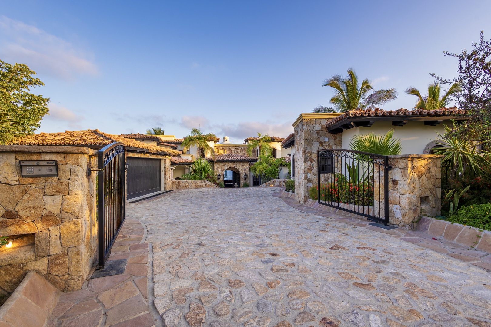 Gated cobblestone entrance to a Spanish-colonial style villa surrounded by lush palms and tropical landscaping under a warm, golden-hour sky.