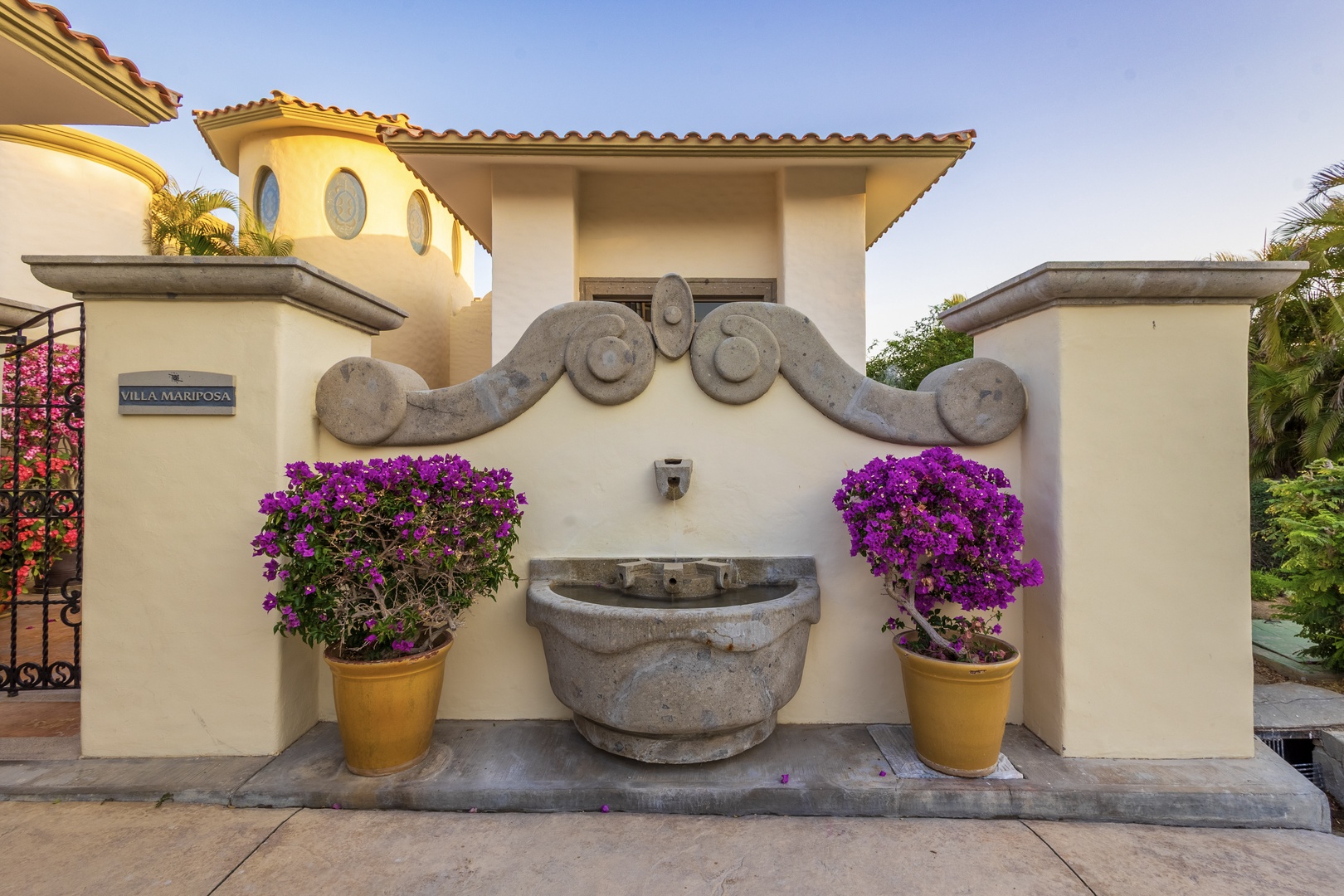 A charming Spanish colonial-style entrance fountain welcomes guests to Villa Mariposa, framed by vibrant purple bougainvillea and terracotta planters.