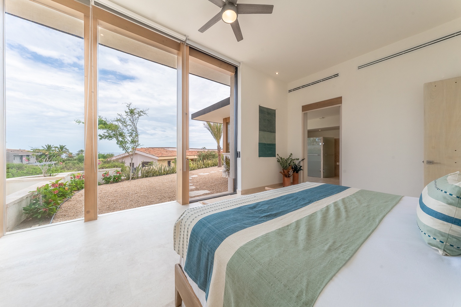 Your serene bedroom opens to a private terrace overlooking lush gardens and distant views. Sink into coastal-inspired linens cooled by the gentle ceiling fan as natural light fills the space.
