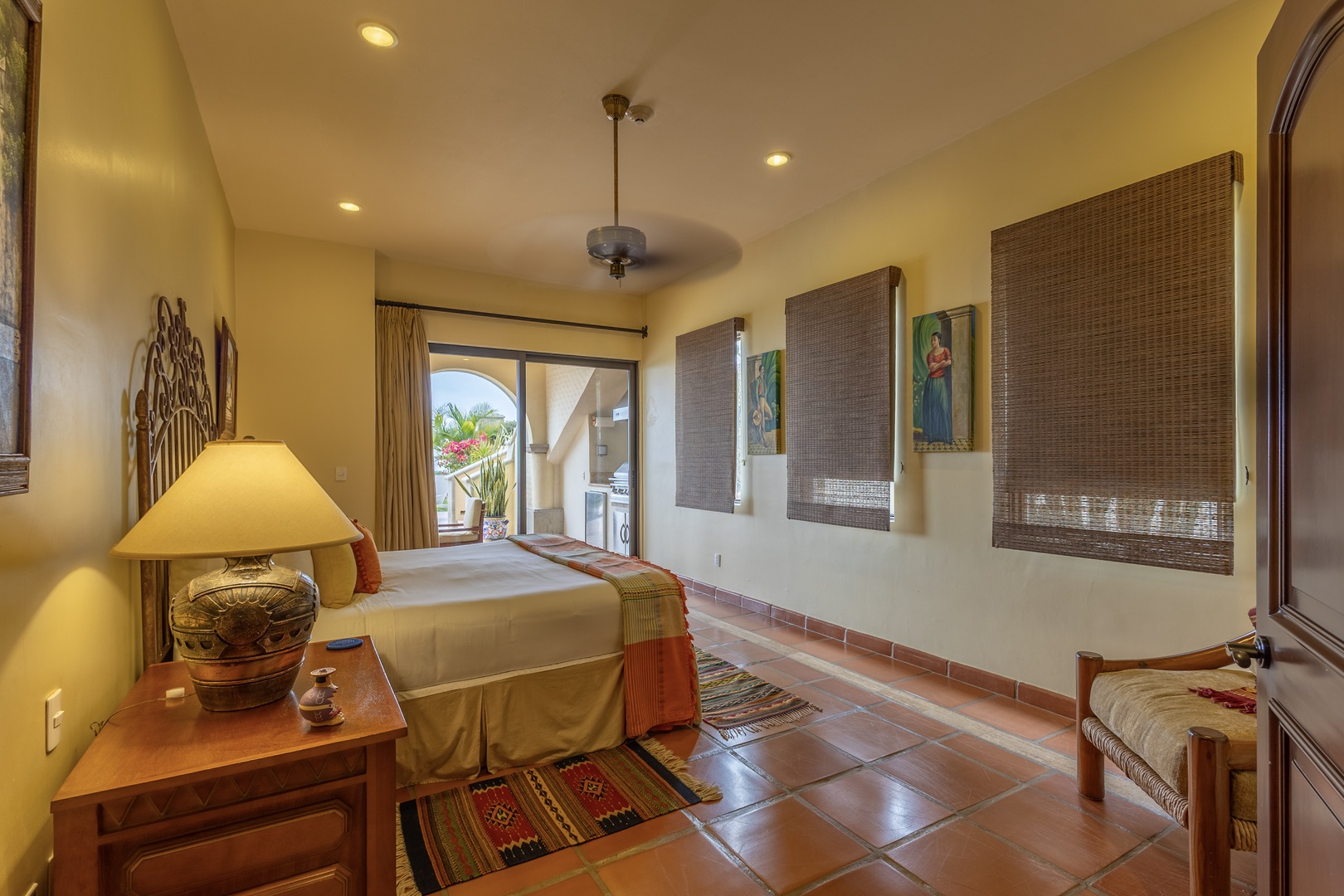 Wake up in your cozy bedroom with warm tile floors, soft natural light filtering through woven shades, and a private balcony doorway framing tropical greenery just beyond.