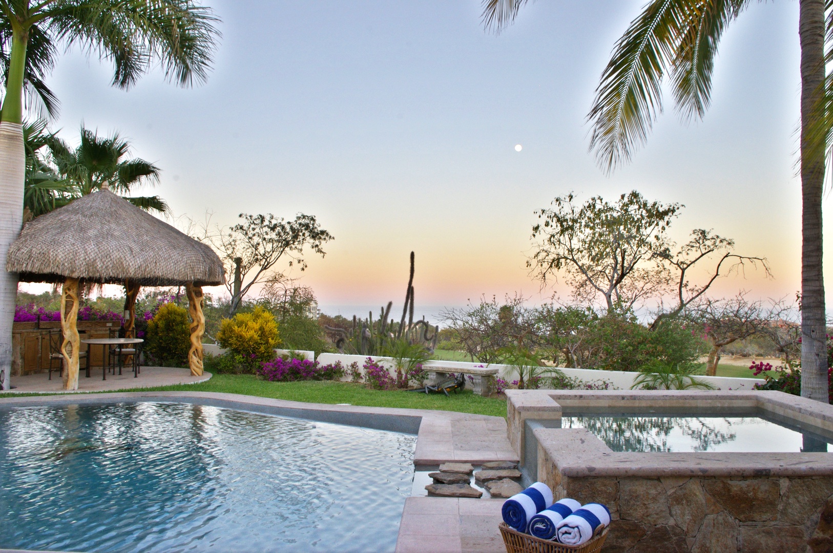 A peaceful desert oasis featuring a sparkling pool, bubbling hot tub, and palapa lounge under soft twilight skies—your private retreat awaits.