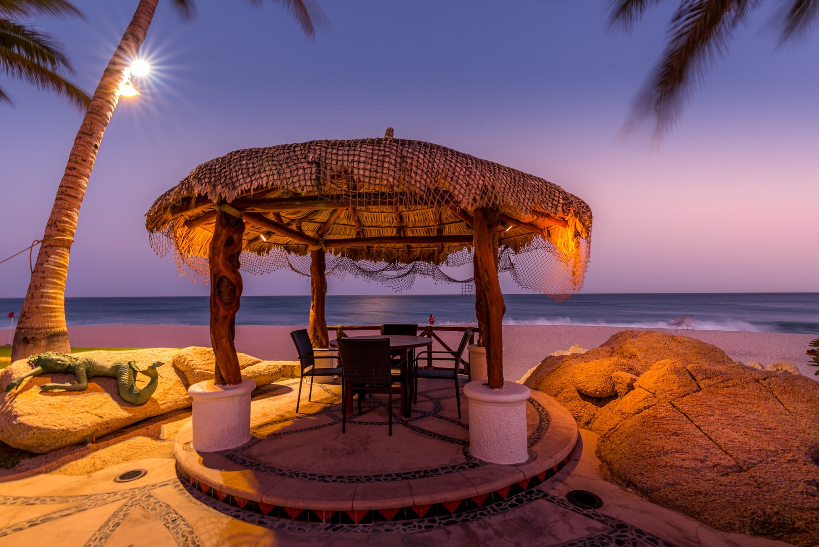 A stunning beachfront dining gazebo awaits at twilight, where the golden glow of evening light meets the gentle ocean waves. Palm trees sway overhead as soft illumination creates a magical, romantic atmosphere perfect for unforgettable seaside moments.