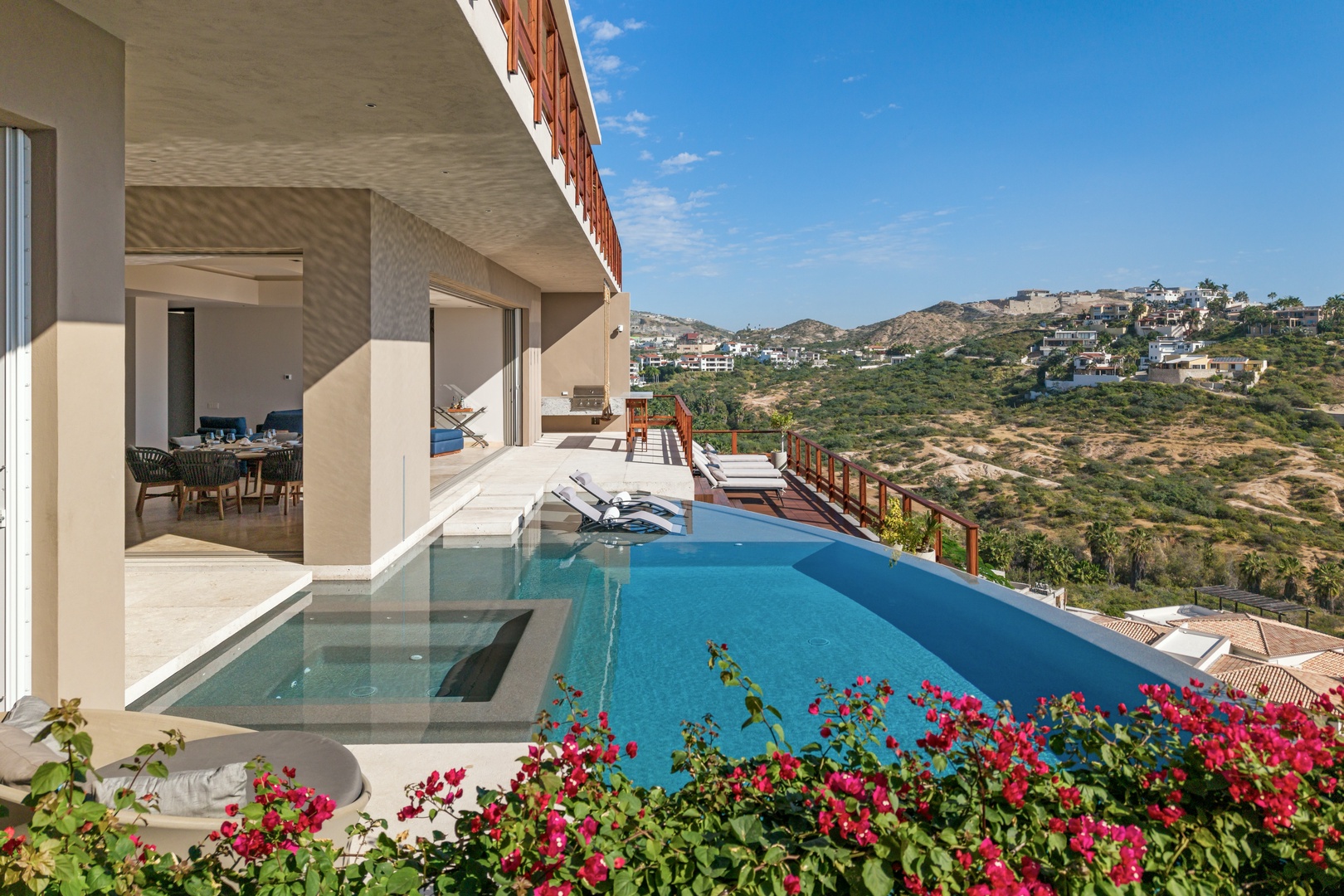 Stunning infinity pool framed by vibrant flowers overlooks rolling hillside vistas. Sunbeds await alongside sleek terrace, blending indoor-outdoor living with breathtaking mountain views under blue skies.