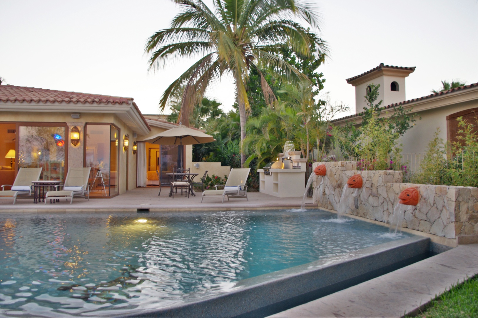 Private Mediterranean-style villa featuring a sparkling pool with decorative water features, lush tropical landscaping, and inviting loungers under swaying palms.