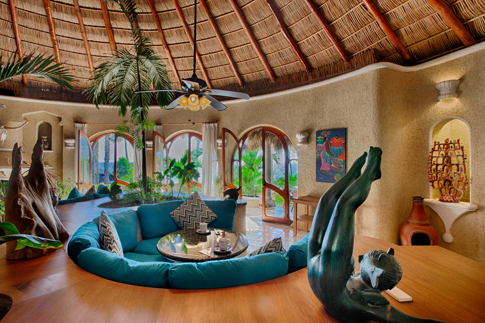 Relax in your stunning circular living room featuring vaulted palm-thatched ceilings, vibrant turquoise seating, and tropical artwork creating an exotic paradise retreat.