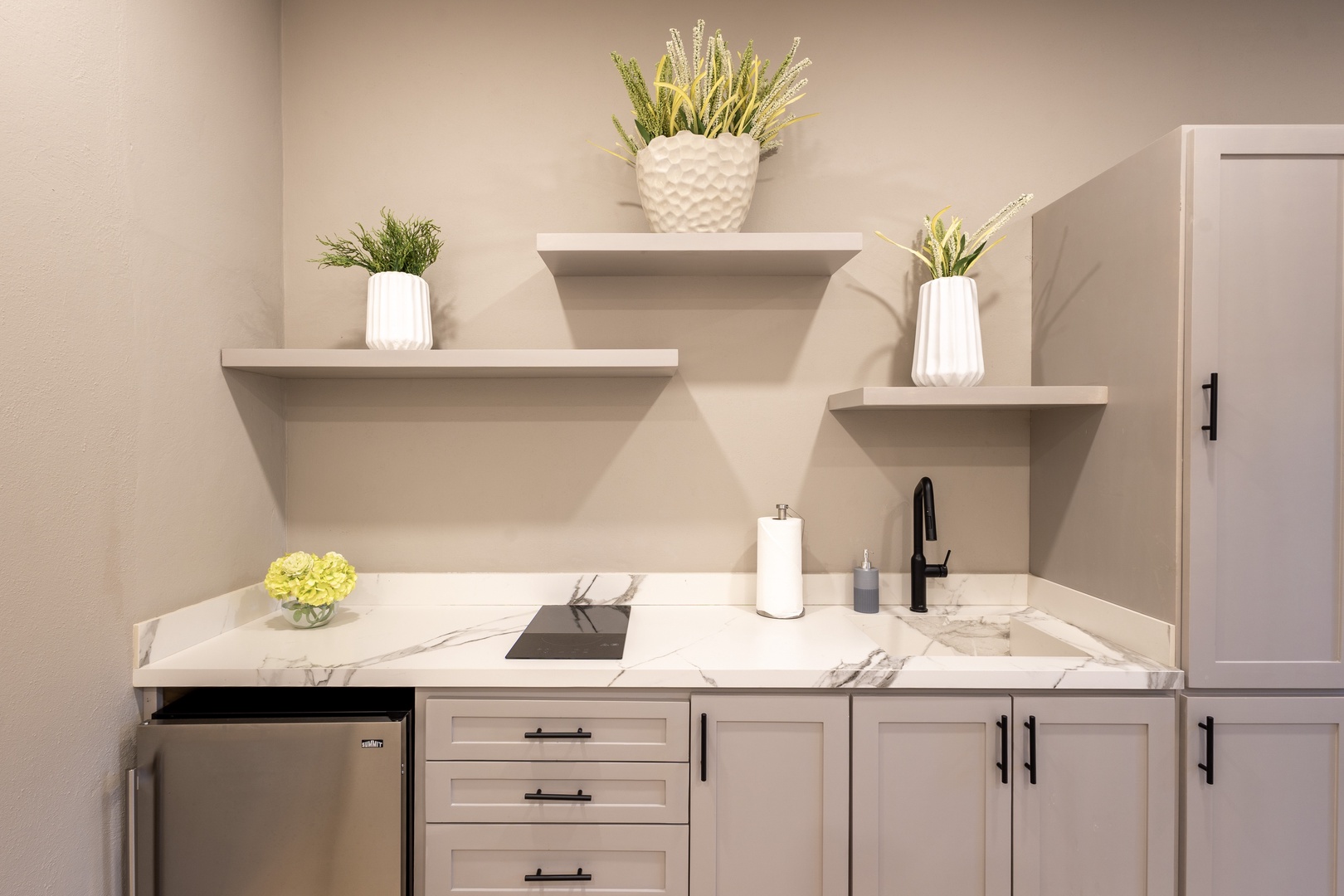 Your private kitchenette features marble-look countertops, modern fixtures, and thoughtful shelving with fresh greenery—a stylish space to prepare light meals or morning coffee.