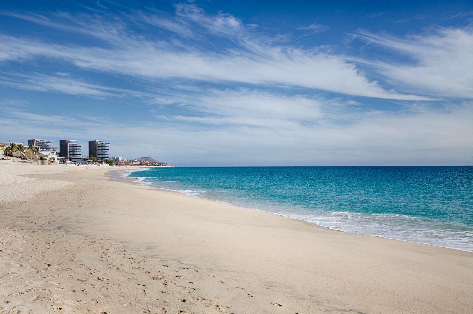 Pristine sandy beach with turquoise waters and coastal development backdrop under brilliant blue skies.