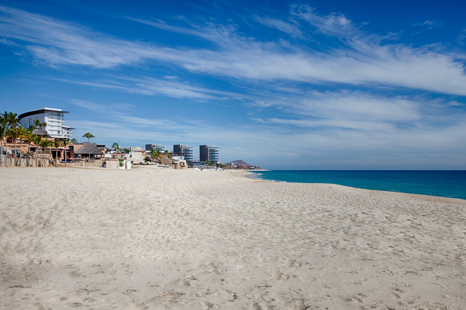 Pristine sandy beach stretches along turquoise waters with modern coastal buildings and palm trees dotting the shoreline.