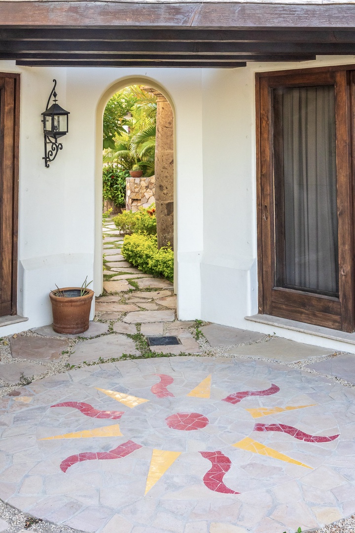 Charming Mediterranean-style entrance features rustic wooden beams, a decorative mosaic floor, and an arched doorway opening to a lush garden courtyard with stepping stones.