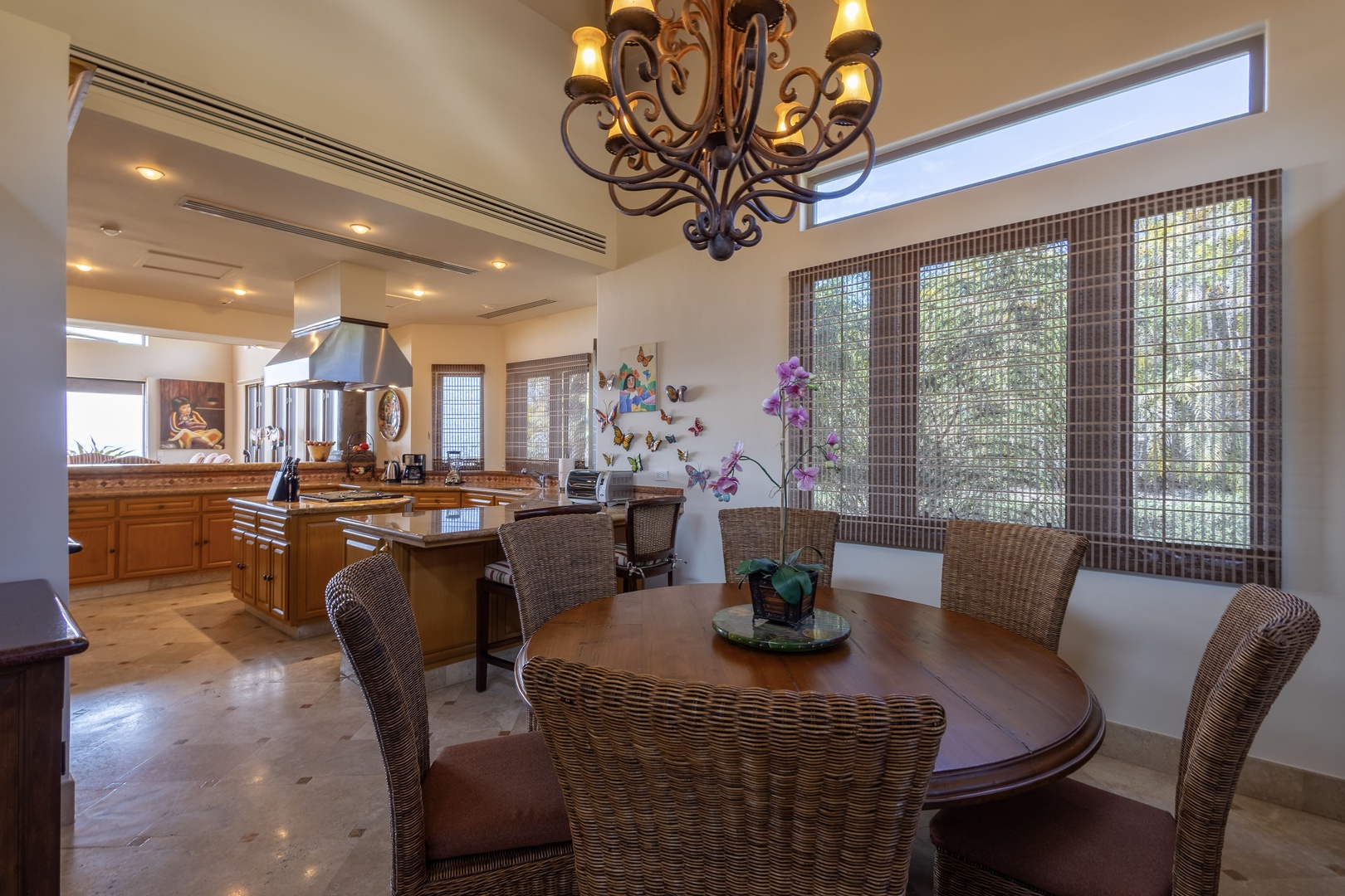 Step into your open-concept kitchen and dining space, where a rustic chandelier casts a warm glow over elegant wicker seating. Large windows frame lush garden views, filling the room with natural light as you prepare meals and gather for dinner.