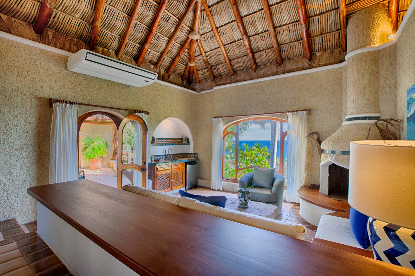 Relax in your villa's open living space featuring traditional thatched ceilings, arched windows with tropical views, and a cozy fireplace for perfect evenings.