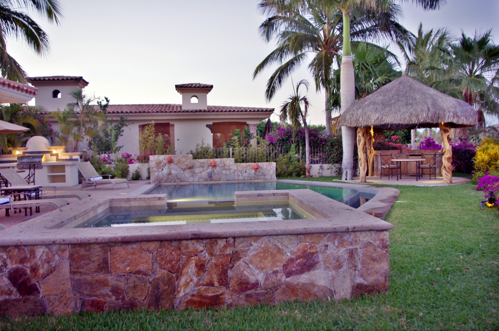 A stunning tropical estate featuring lush gardens, sparkling pool, and thatched palapa dining area surrounded by vibrant bougainvillea and towering palms.