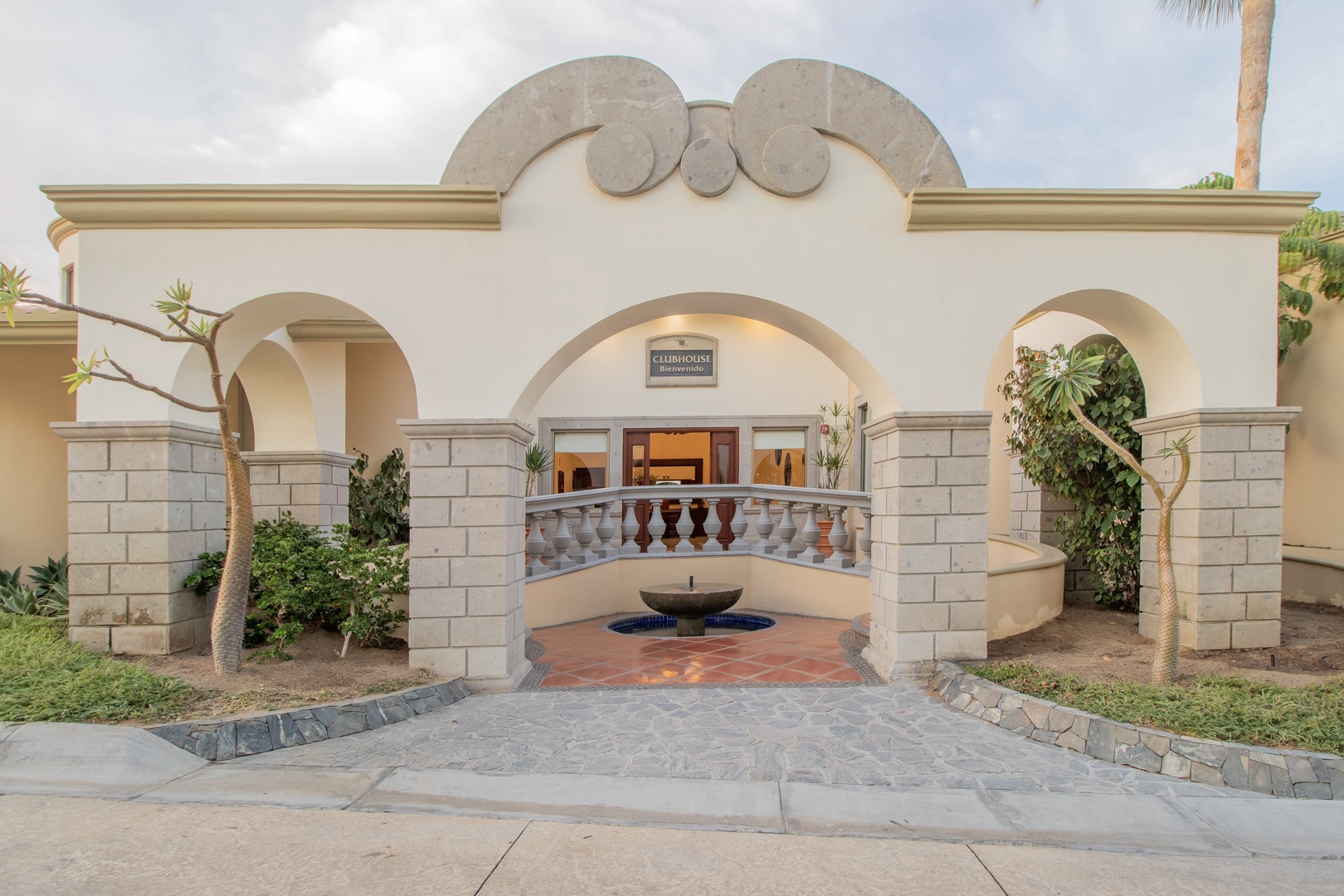 Mediterranean-inspired entrance welcomes visitors with elegant archways, decorative stonework, and a tranquil fountain centerpiece within a landscaped courtyard setting.