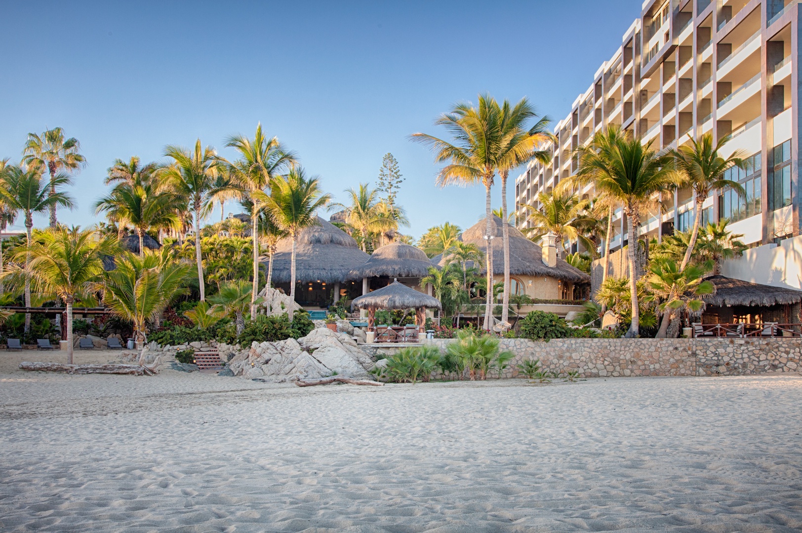 Beachfront resort property featuring tropical architecture with thatched-roof buildings, swaying palm trees, and direct beach access under clear blue skies.