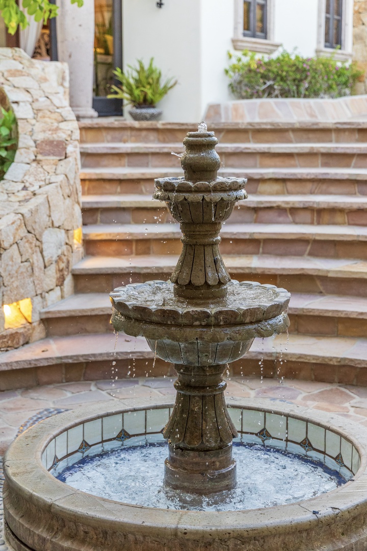 A tiered stone fountain anchors the property's entryway courtyard, surrounded by Mediterranean-style landscaping, warm stonework, and natural architectural details leading to the main entrance.