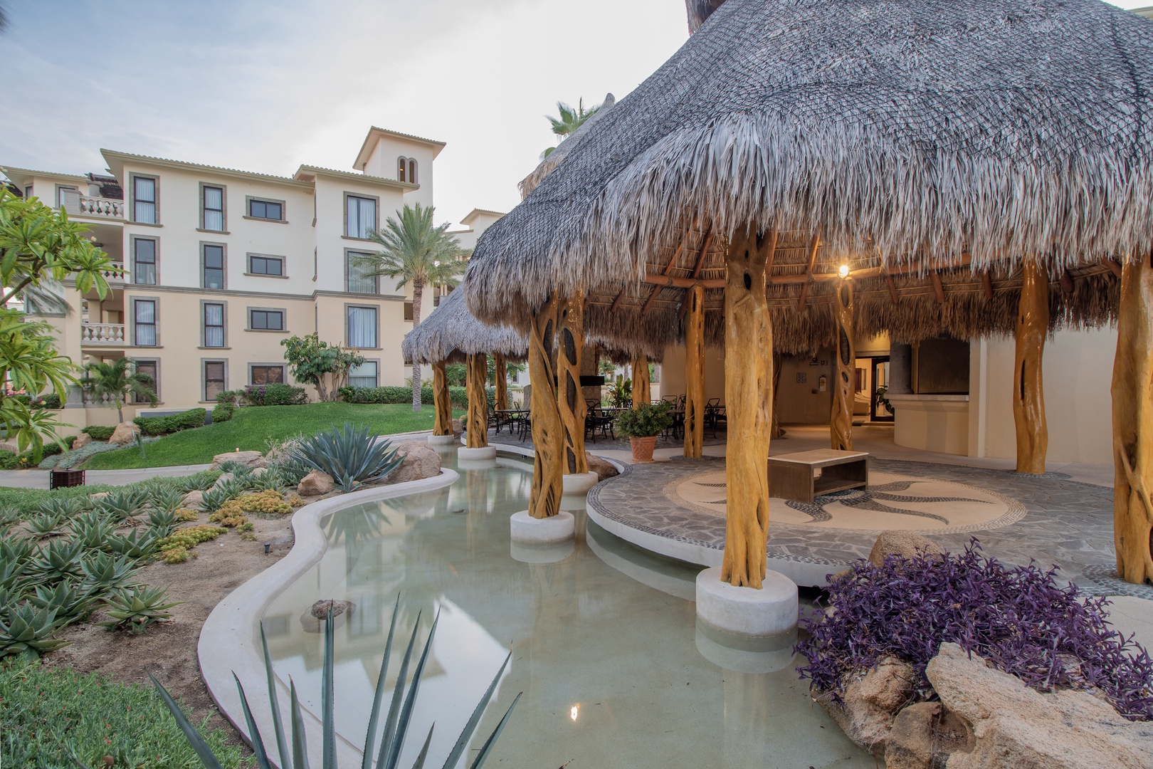 A serene property garden features a tranquil water feature and tropical palapa structure, surrounded by lush landscaping, native plants, and well-maintained lawns—creating a peaceful retreat setting.