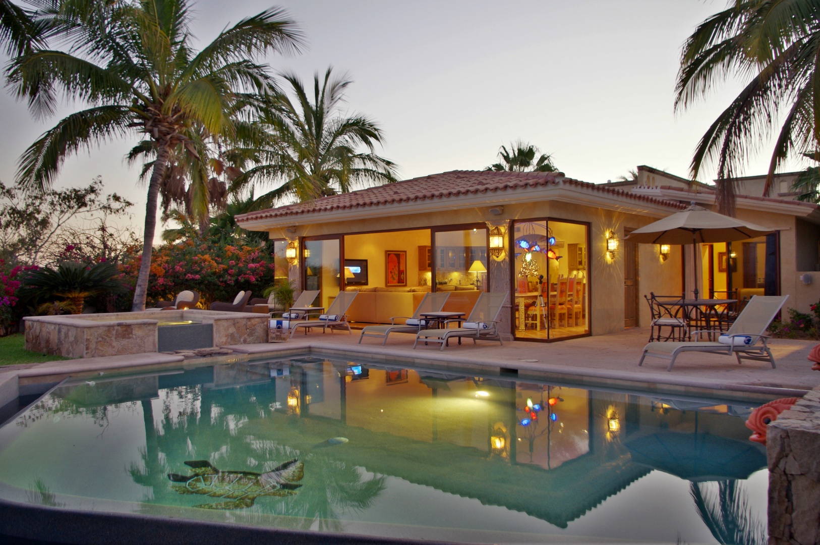 A stunning Spanish-style villa surrounded by tropical palms, featuring a sparkling pool that perfectly reflects the warm interior glow at twilight.