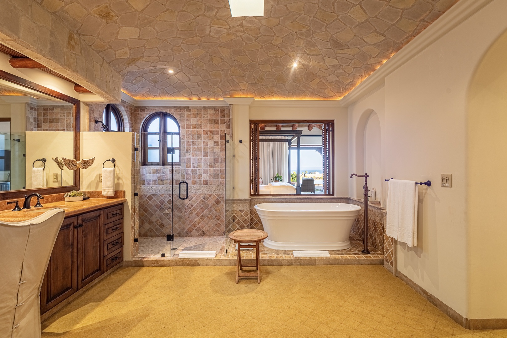 Soak away your cares in this stunning spa-like bathroom featuring a freestanding soaking tub, spacious walk-in shower with intricate tilework, and handcrafted vanity under a vaulted stone ceiling.