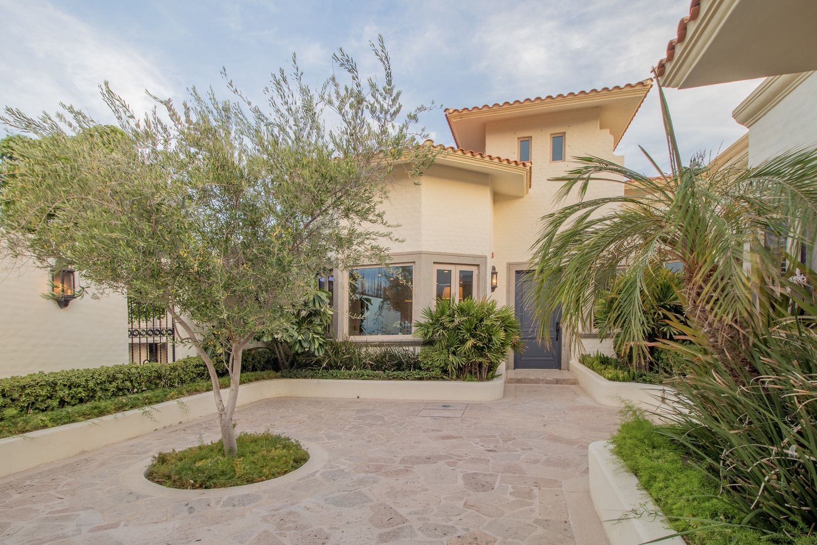 Mediterranean-style villa entrance featuring Spanish tile roof, landscaped driveway with palm trees, and warm stucco exterior, offering an inviting arrival experience.