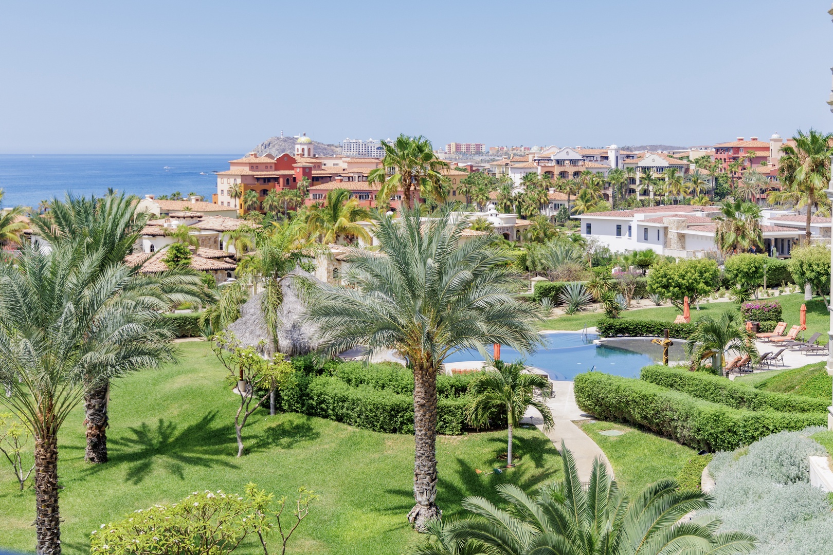 Elevated view capturing the resort's lush tropical gardens, sparkling pool, and the azure sea stretching toward distant coastal cityscape under clear blue skies.