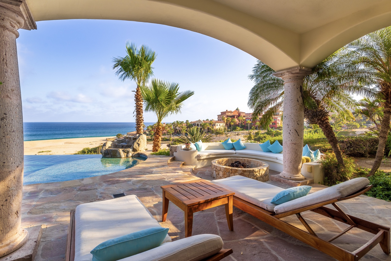 A covered patio frames an infinity pool, beach access, and ocean views framed by swaying palms. Lounge on cushioned chairs by the fire pit and soak in the coastal serenity.