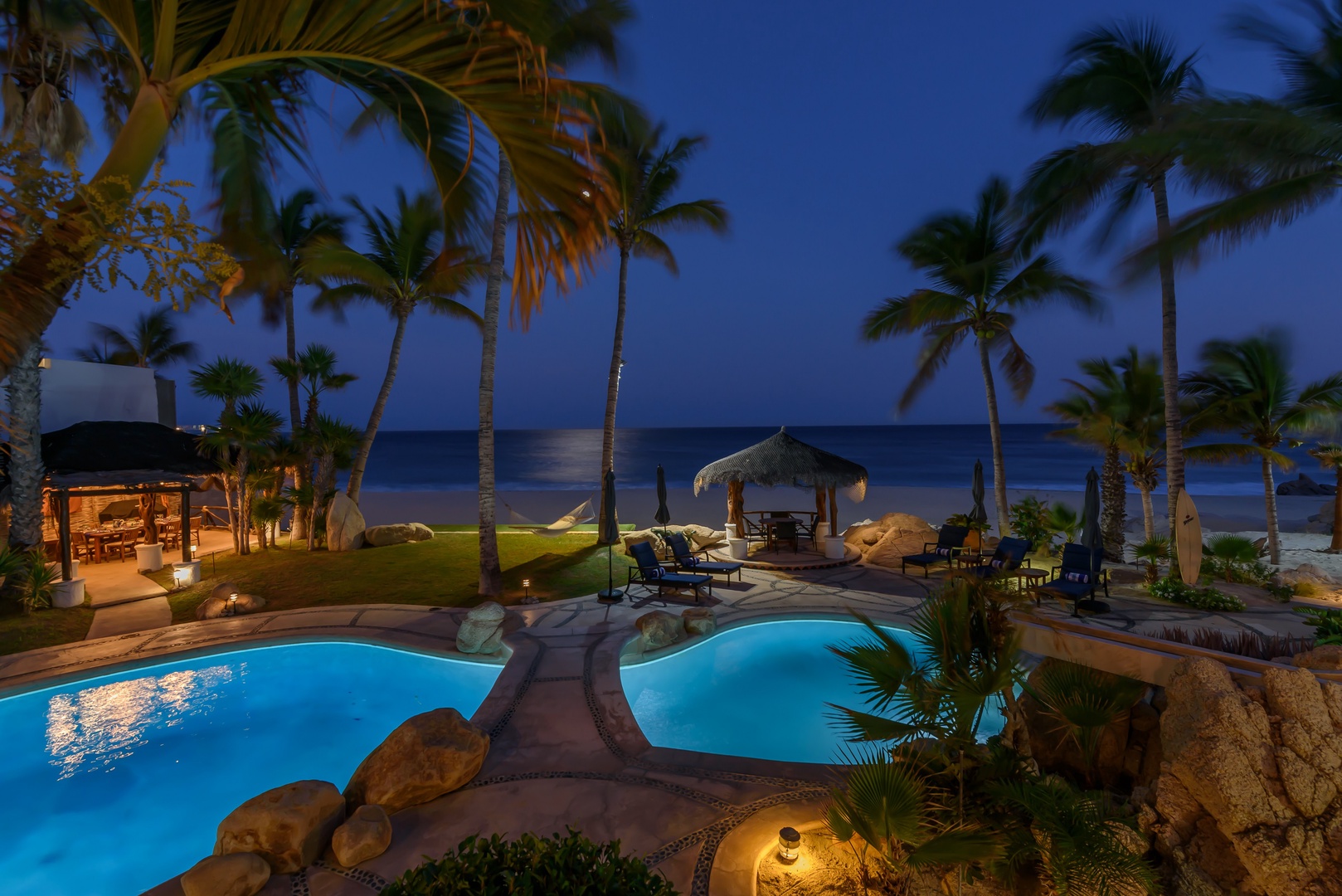 Luxurious beachfront oasis with a stunning pool, thatched palapa, and swaying palms—all set against the tranquil sea at twilight, perfect for an unforgettable tropical escape.