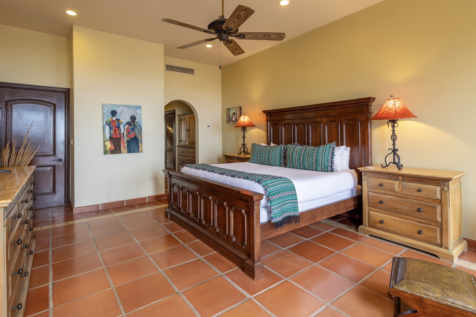 Unwind in your spacious bedroom featuring a beautifully carved wooden bed dressed in crisp linens and teal accents, warm terracotta tile floors, and a cozy ceiling fan to keep you comfortable.